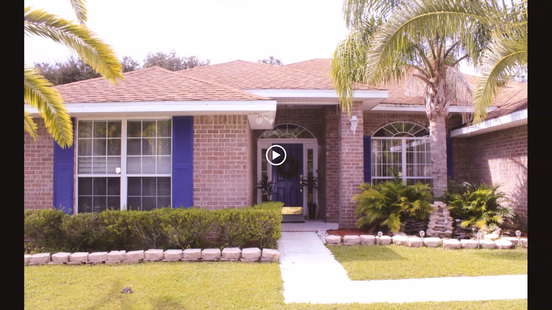 Front exterior view of a single-story brick building with a brown shingled roof, blue shutters on the windows, a purple front door with a wreath, surrounded by green bushes, palm trees, and a well-maintained lawn with a concrete walkway leading to the entrance.