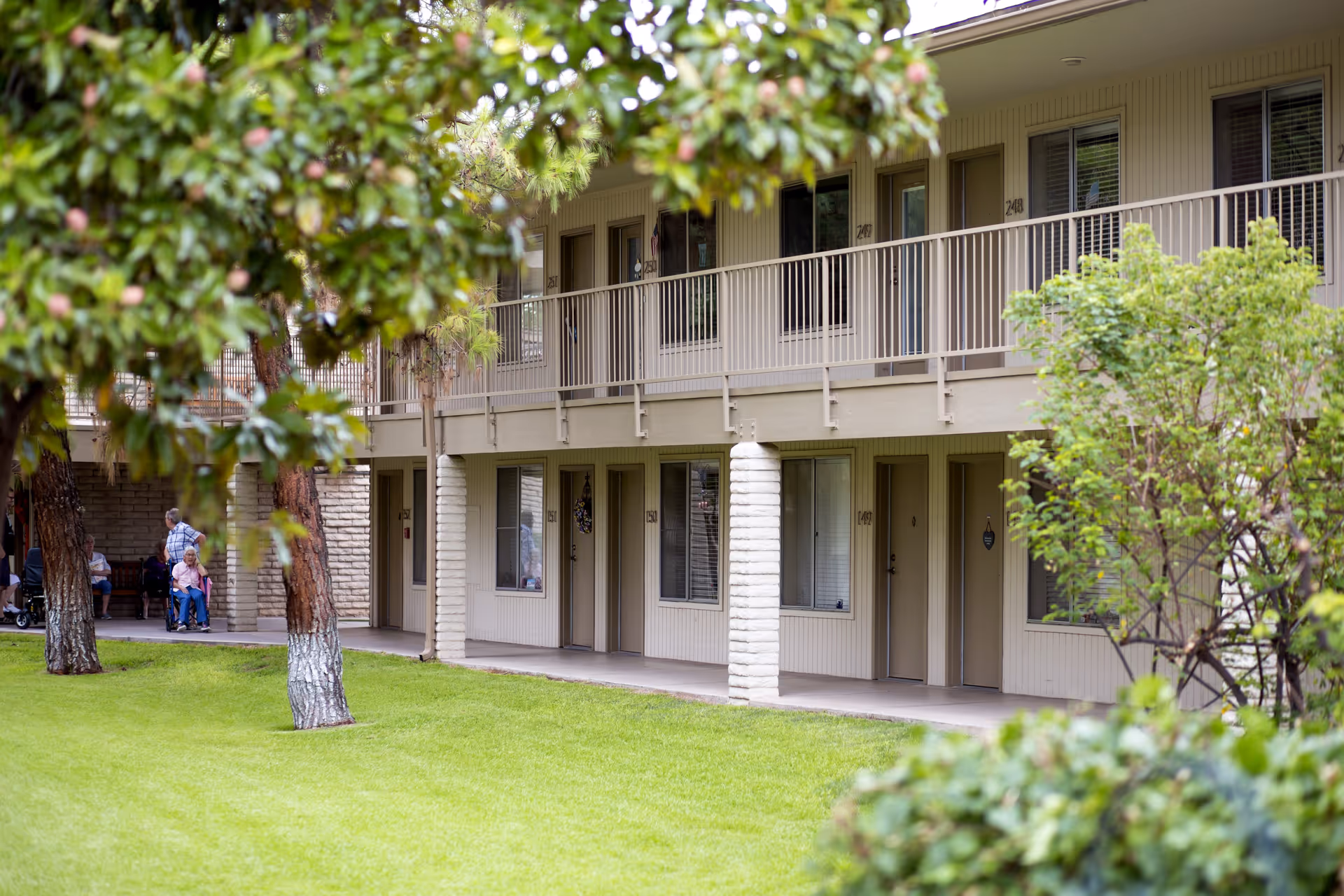 Two-story beige senior living building with balconies, doors, and a green lawn.