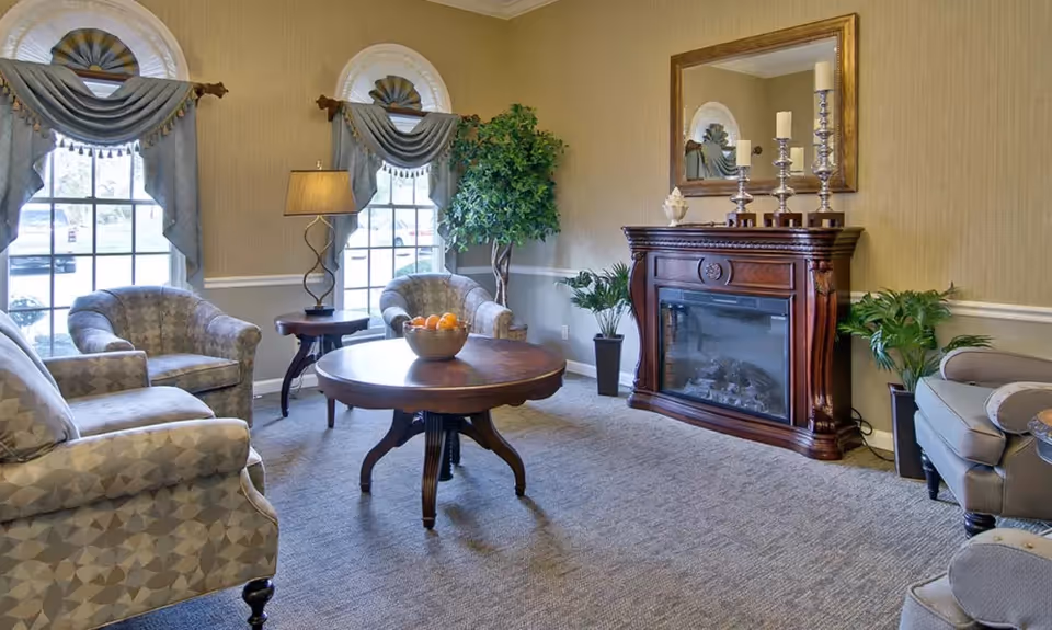 A cozy living room with patterned armchairs, a round wooden coffee table with a bowl of fruit, a wooden electric fireplace with decorative candles and a mirror above it, two large windows with blue valance curtains, a floor lamp, and several potted plants.
