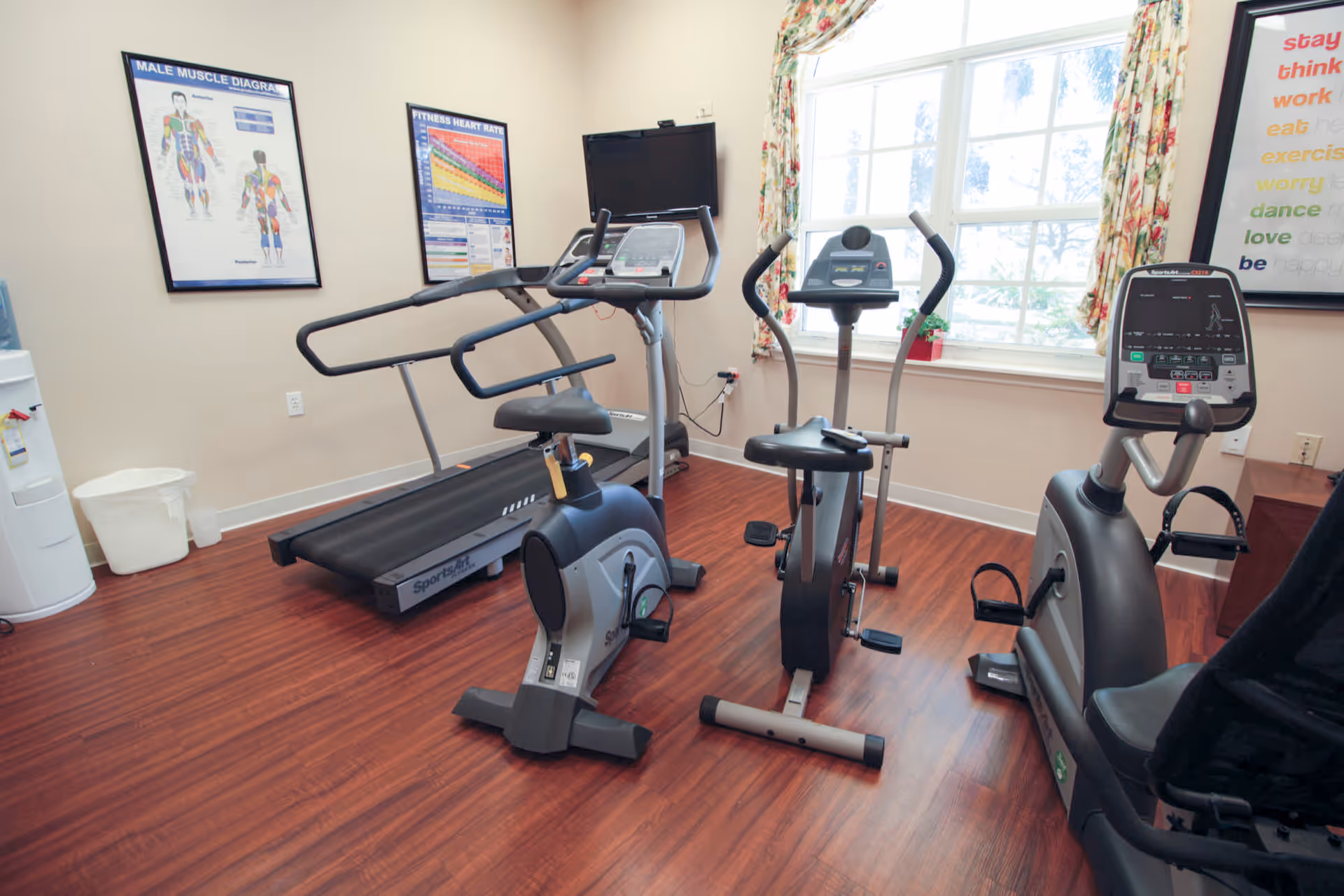 A small fitness room with exercise equipment including a treadmill, two stationary bikes, and an elliptical machine. The room has wooden flooring, a window with floral curtains, and posters on the walls showing a male muscle diagram and fitness heart rate chart. A water cooler and trash bin are visible in the corner.