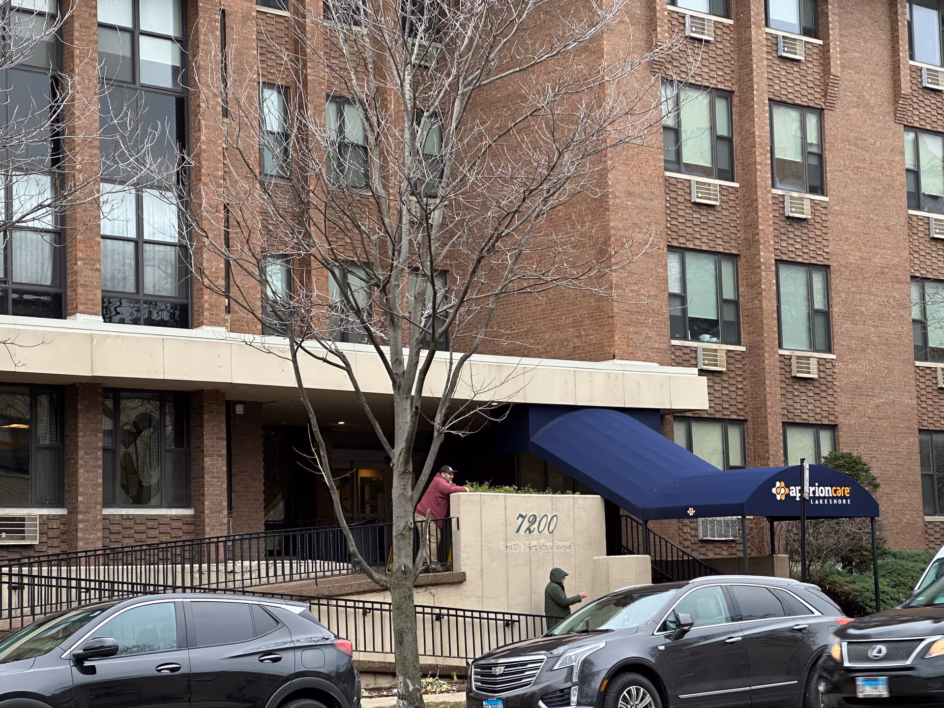 Exterior view of a multi-story brick building with several windows and air conditioning units. A blue awning with the text 'aperion care LAKESHORE' covers the entrance. Two people are visible near the entrance, one standing on a ramp and another walking nearby. Several parked cars are in front of the building, and a leafless tree is in the foreground.