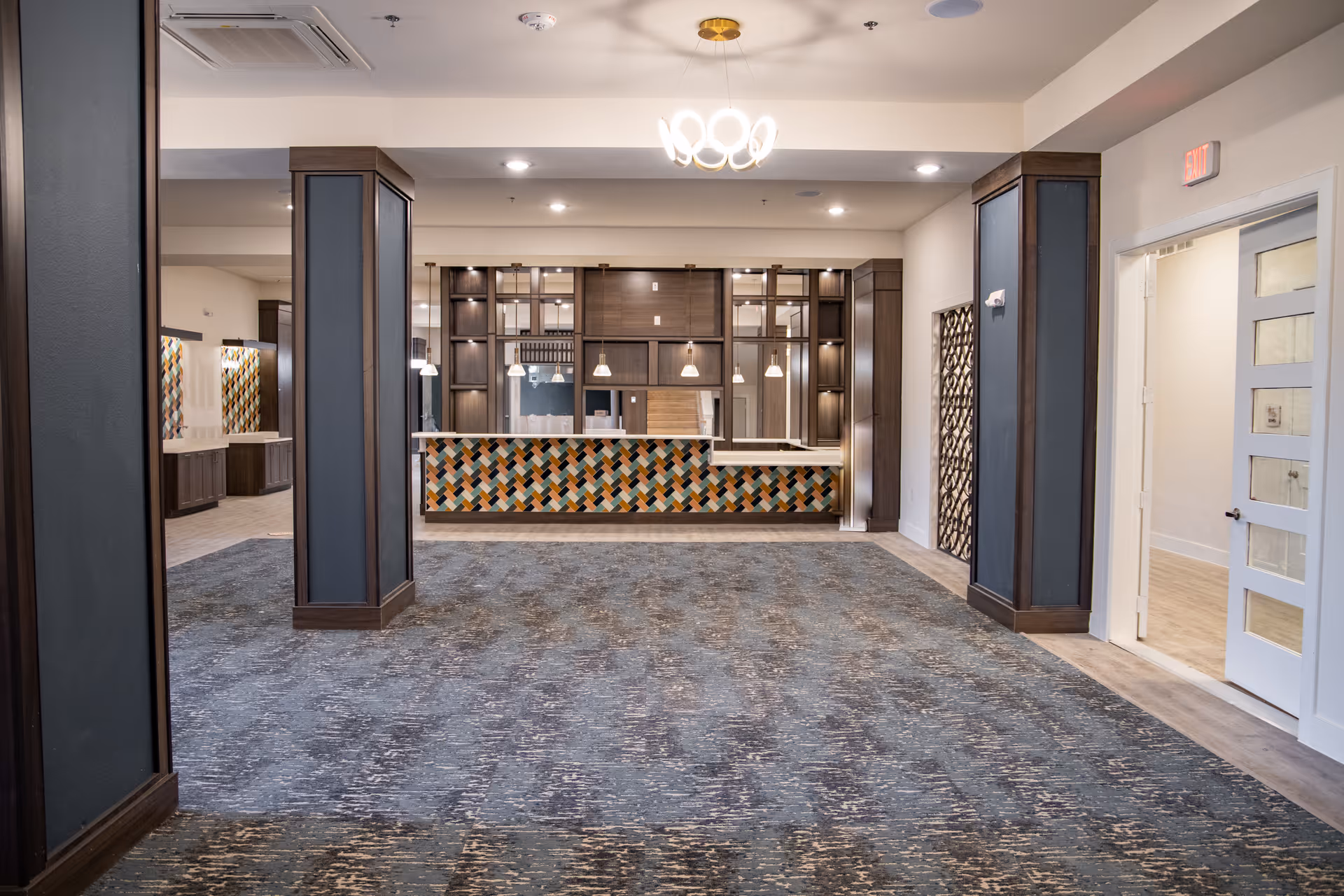Interior view of a spacious room with patterned carpet flooring, two large pillars with dark wood and blue panels, and a reception or service counter with a colorful geometric tile front. The room is well-lit with ceiling lights and a modern chandelier. There is a door on the right side leading to another room with light wood flooring.
