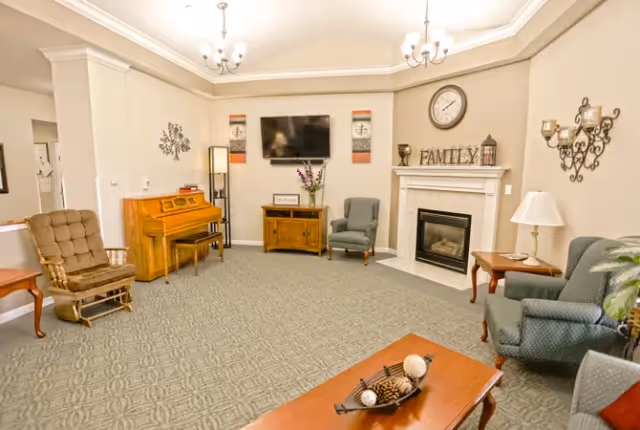 Communal living room with a fireplace, wall-mounted TV, piano, armchairs, and a coffee table.