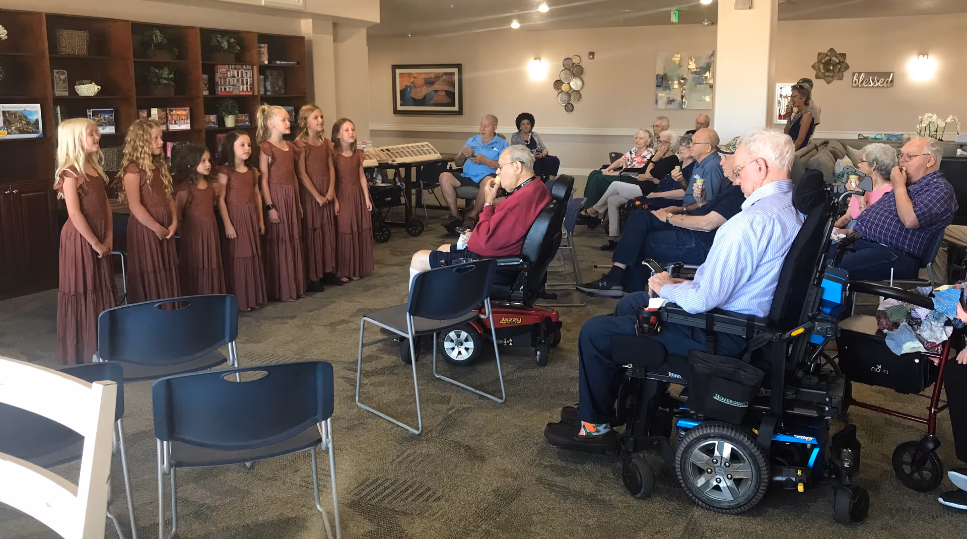 A group of young girls in matching brown dresses standing and singing in front of an audience of elderly people seated in chairs and wheelchairs in a common room with bookshelves and wall decorations.