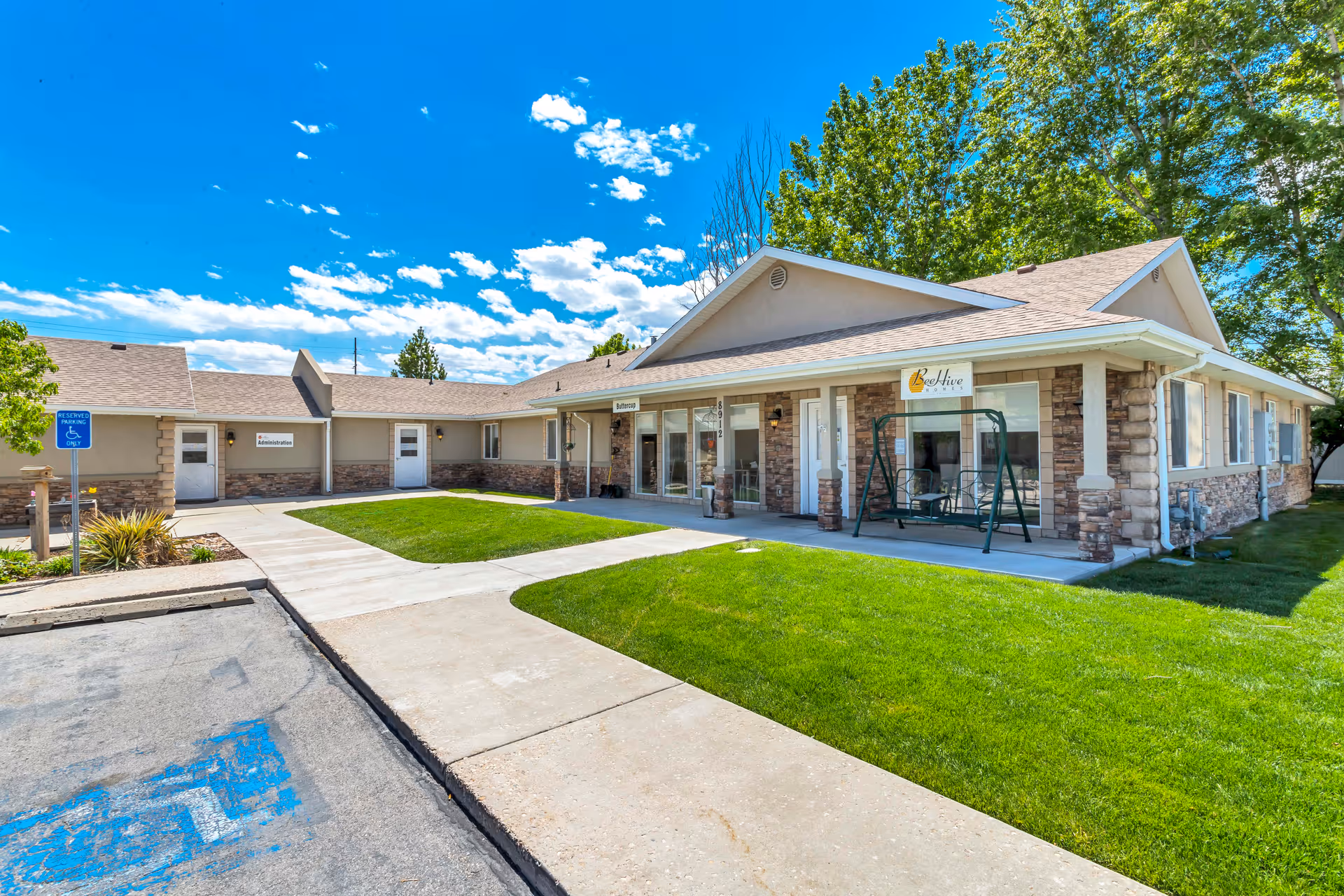 Exterior view of a single-story senior living facility building with beige walls and stone accents. There is a concrete walkway leading to the entrance, green grass lawns, a green porch swing, and signs indicating 'Administration' and 'BeeHive Homes'. The sky is blue with scattered clouds and there are trees in the background.