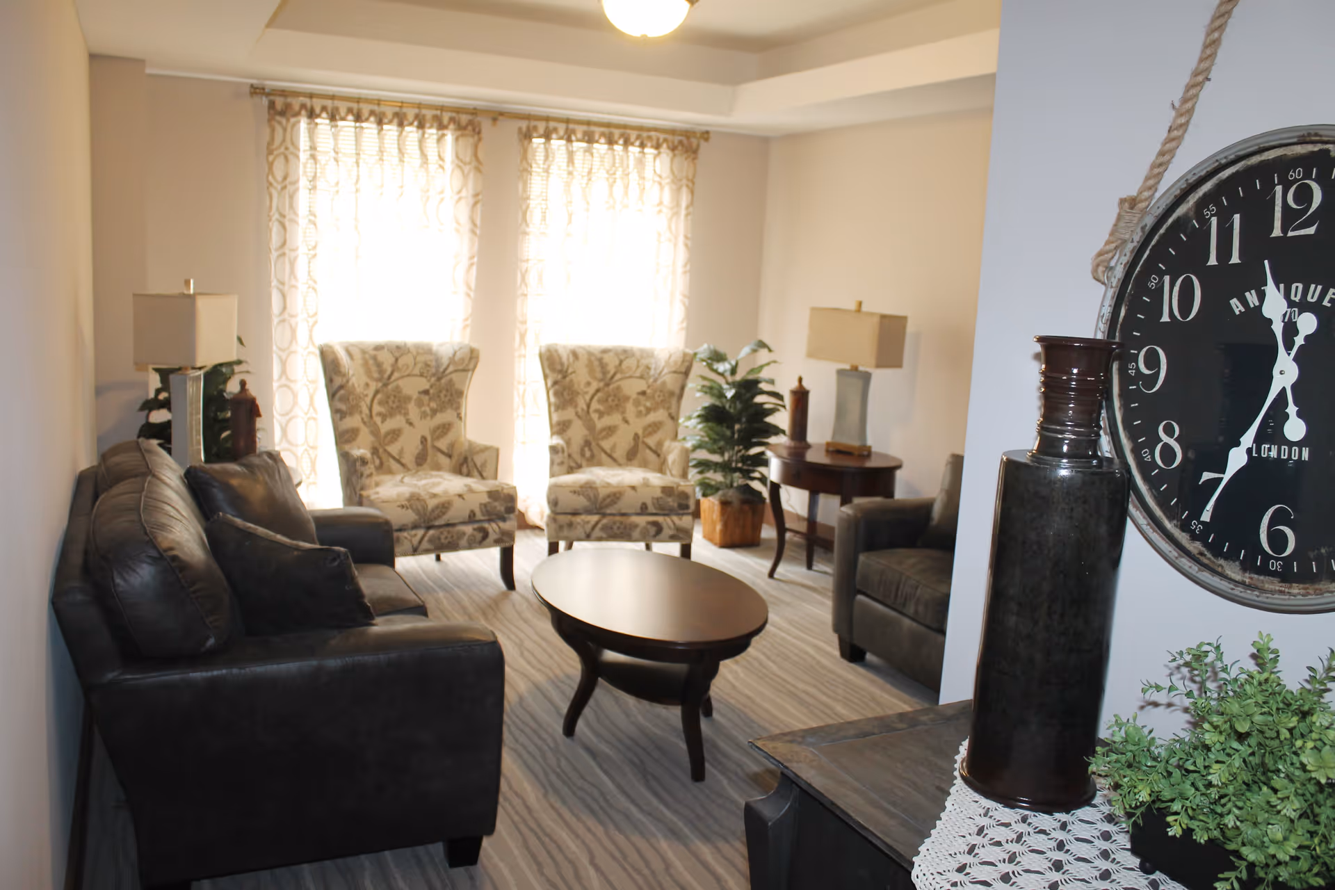 A cozy living room with a dark leather sofa, two patterned armchairs, a dark armchair, and a wooden oval coffee table. The room has light-colored walls, two windows with sheer patterned curtains, two side tables with lamps, a potted plant, and a large wall clock hanging on a partial wall near a decorative vase and greenery on a wooden surface.