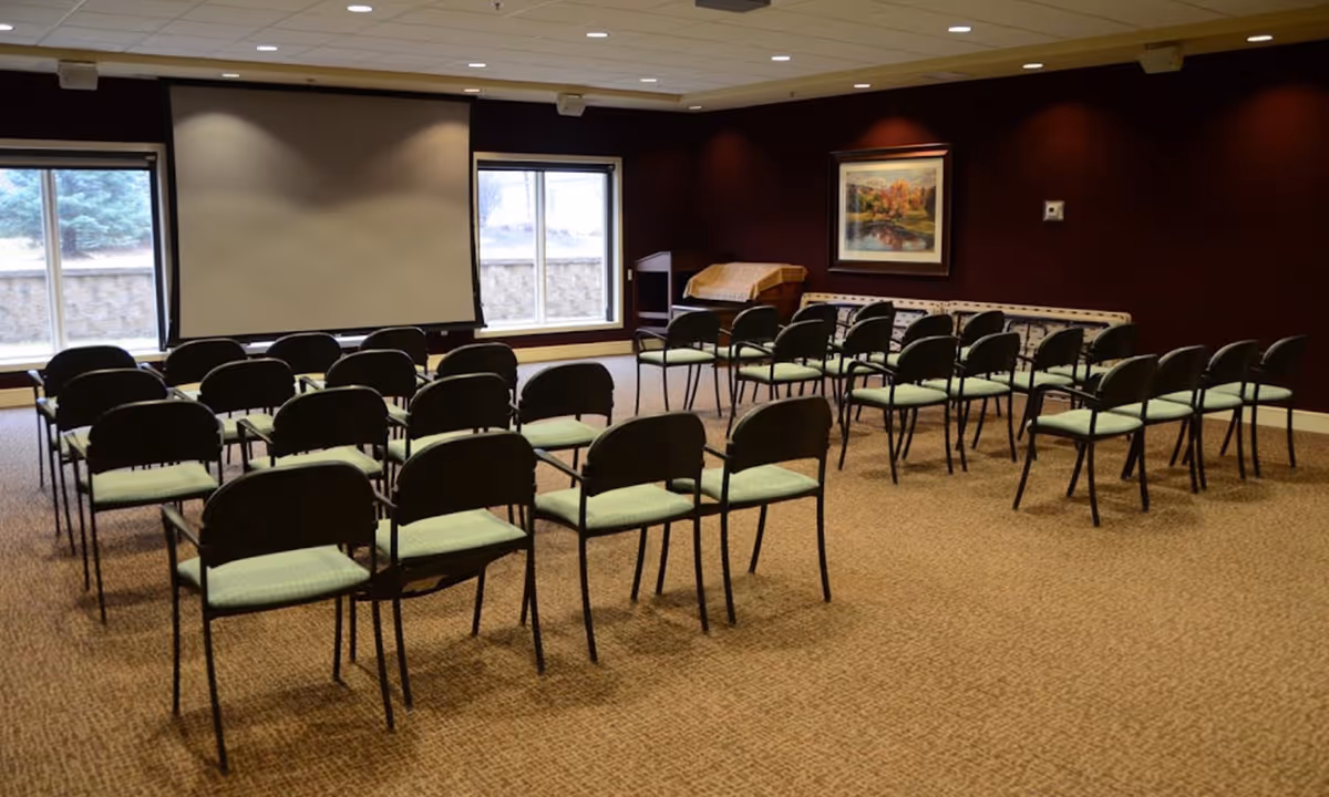 Carpeted meeting room with rows of chairs facing a large projection screen and windows.