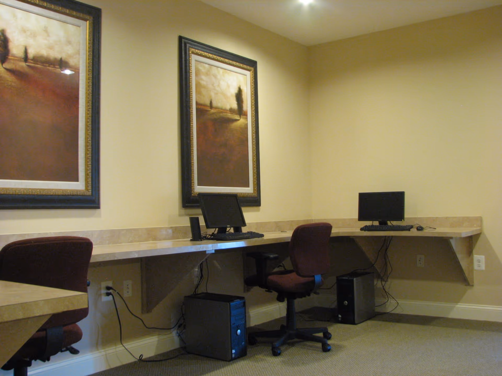 A small computer workstation area with two desktop computers on a beige countertop. Two maroon office chairs are positioned in front of the computers. The walls are painted light beige and decorated with two framed landscape paintings. The floor is carpeted in a neutral tone.