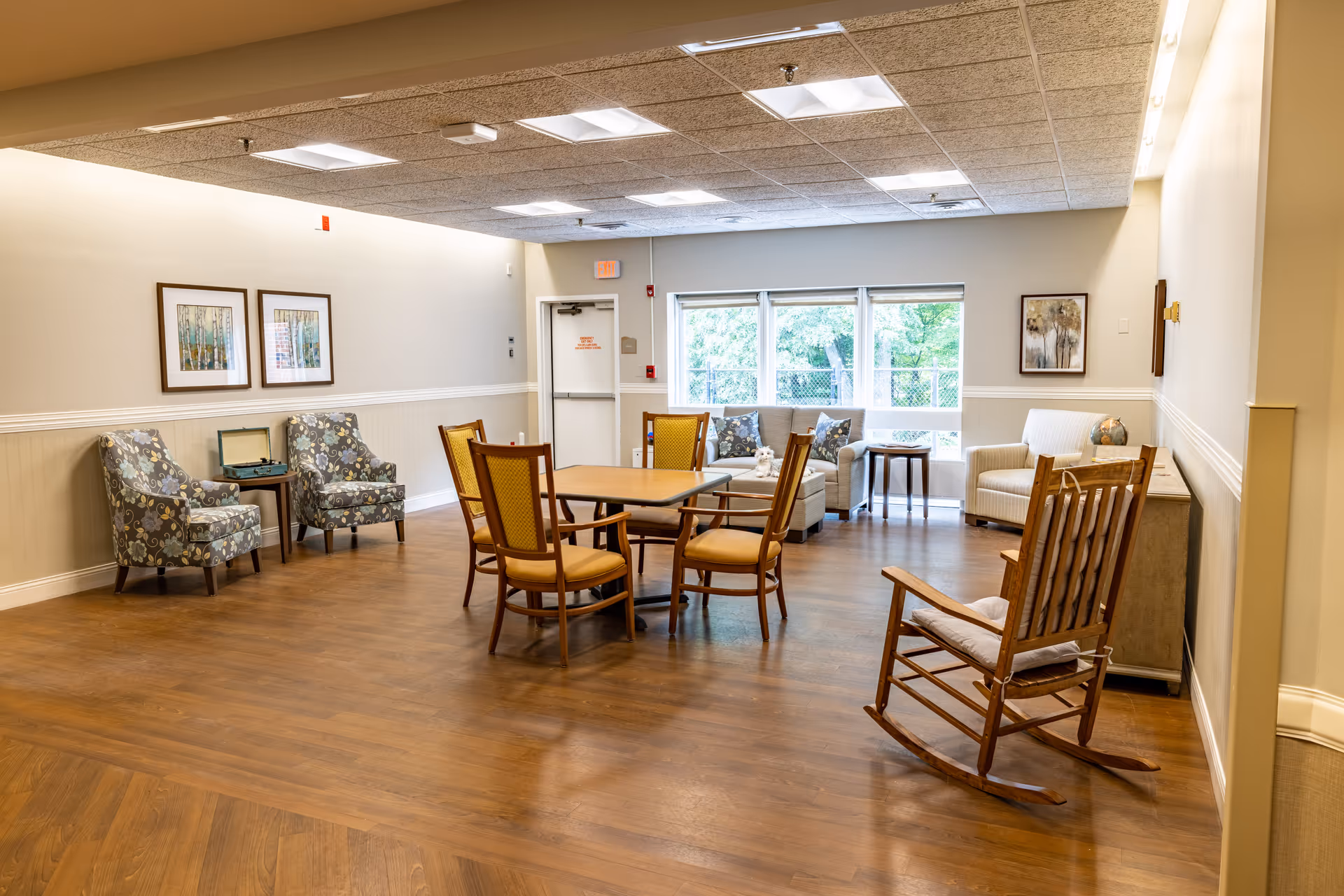 A bright communal living room with wooden floors, armchairs, a round table with chairs, and a wooden rocking chair near large windows.
