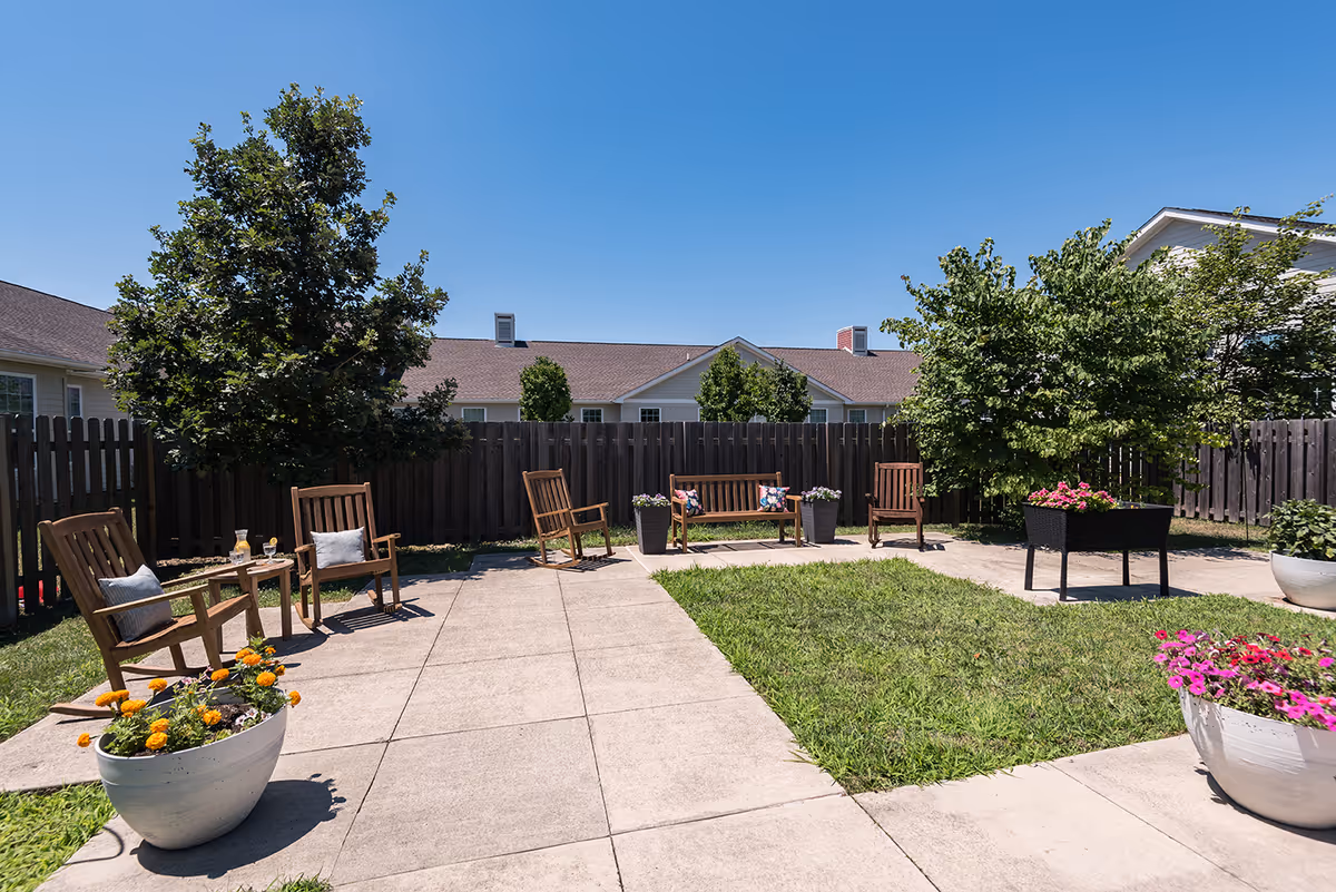 Sunny fenced courtyard with wooden chairs and benches, potted flowers, grass, and a paved walkway.
