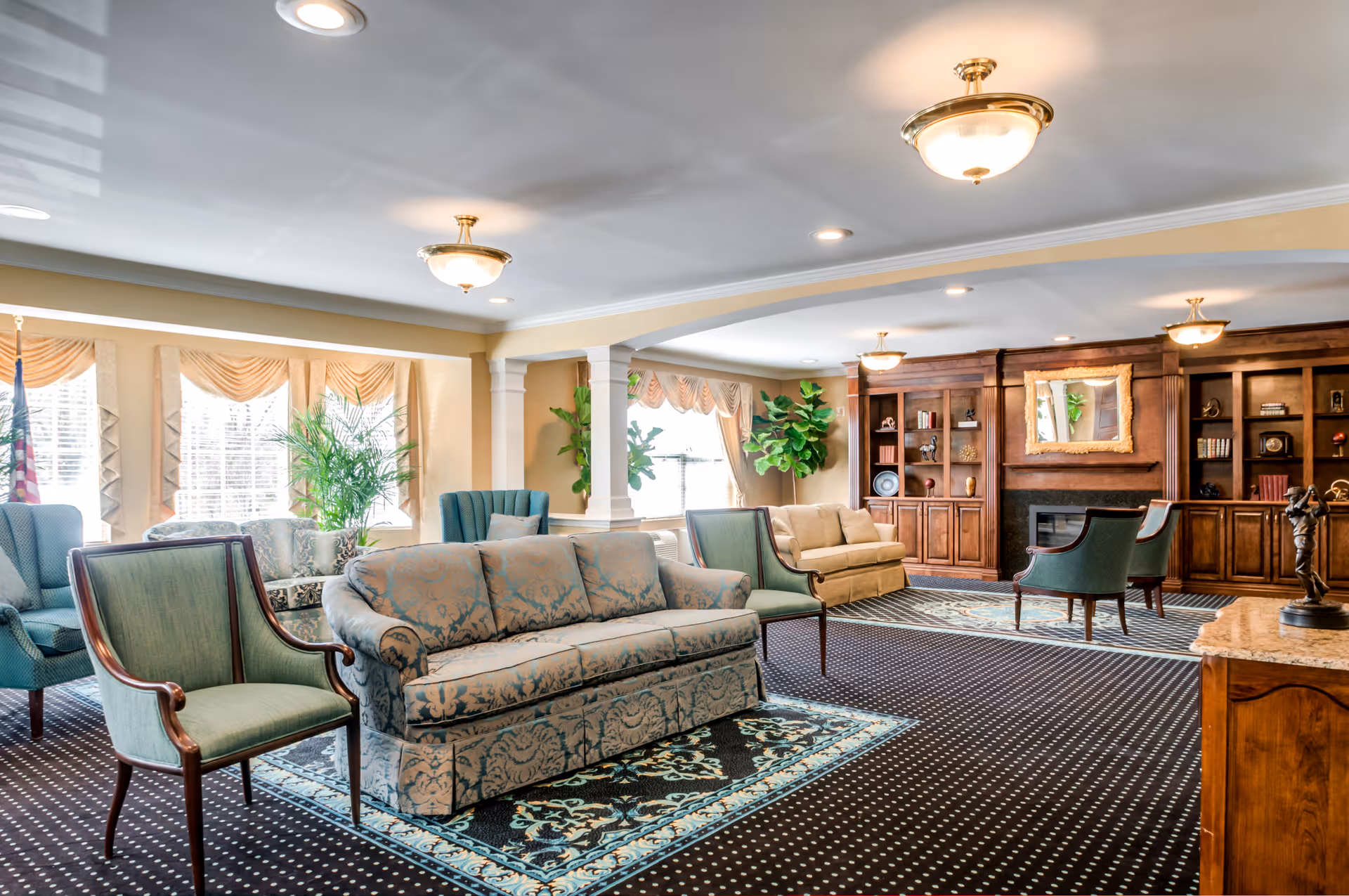 A spacious and well-lit living room area with patterned sofas and armchairs arranged on decorative rugs. Large windows with draped curtains allow natural light to fill the room. The back wall features built-in wooden shelves and cabinets surrounding a fireplace with a framed mirror above it. Potted plants add greenery to the space.