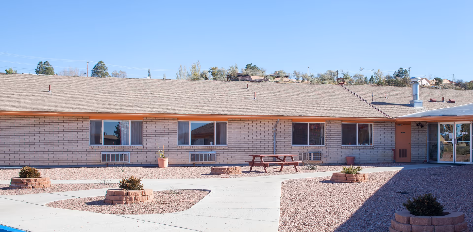 Single-story brick nursing facility front with windows, a picnic table, and low brick planters in a gravel courtyard.