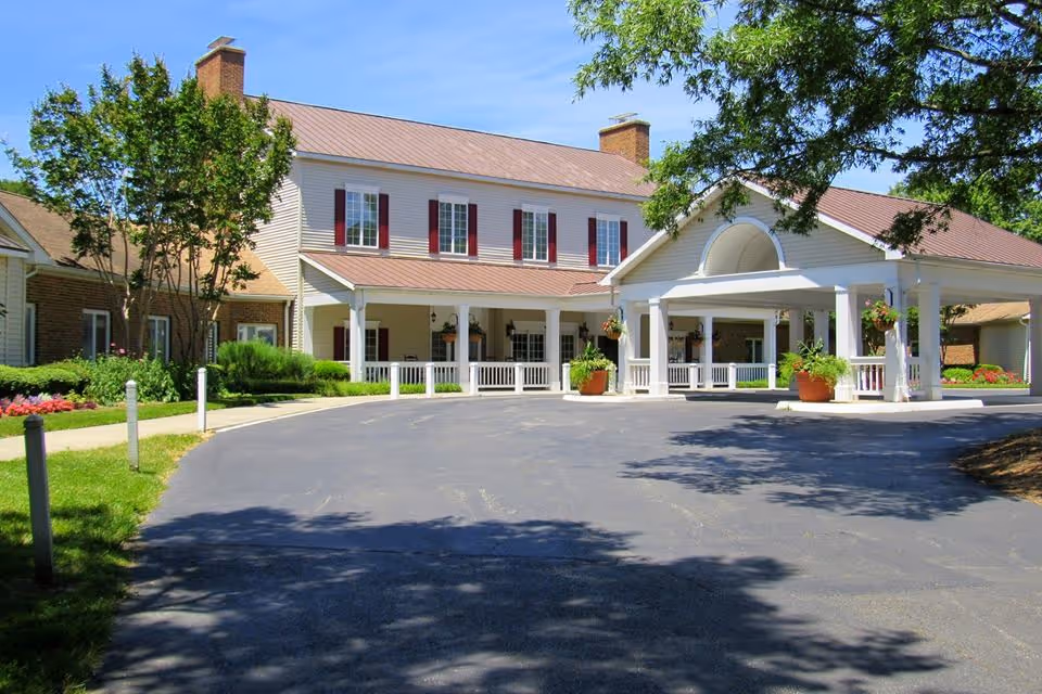 Front entrance of a two-story senior living building with a covered porte-cochere, driveway, and landscaped grounds.