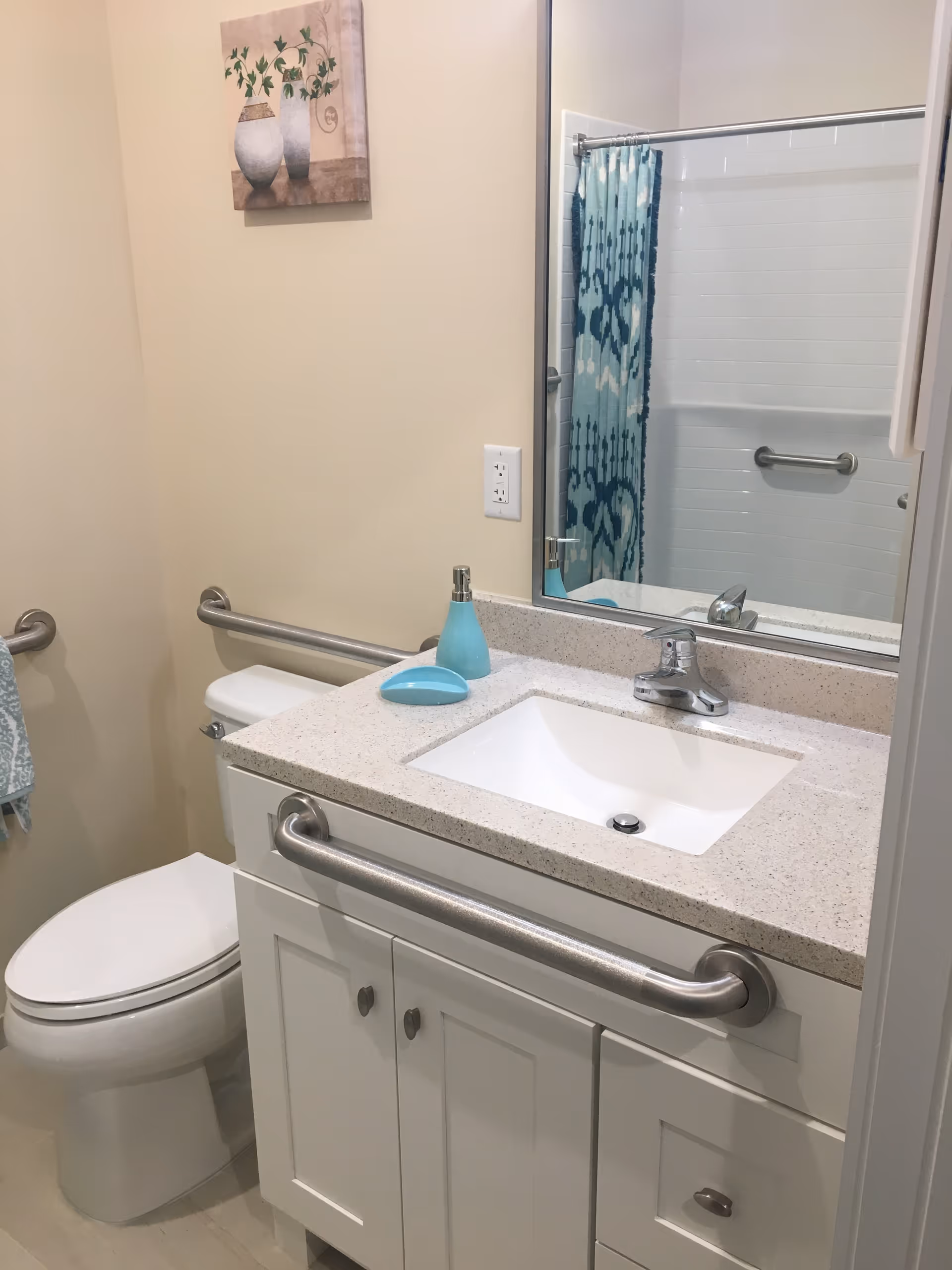 A clean bathroom with a white toilet, a white vanity with a beige countertop, a sink, and a silver faucet. There are stainless steel grab bars on the vanity and next to the toilet. A blue soap dispenser and soap dish are on the countertop. A large mirror is above the sink, reflecting a shower with a blue patterned curtain and a grab bar. A small painting of vases with green plants hangs on the wall.