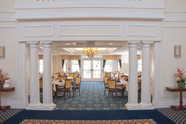 Elegant dining room with multiple set tables and chairs, a chandelier, and white columns framing the entrance over a blue patterned carpet.