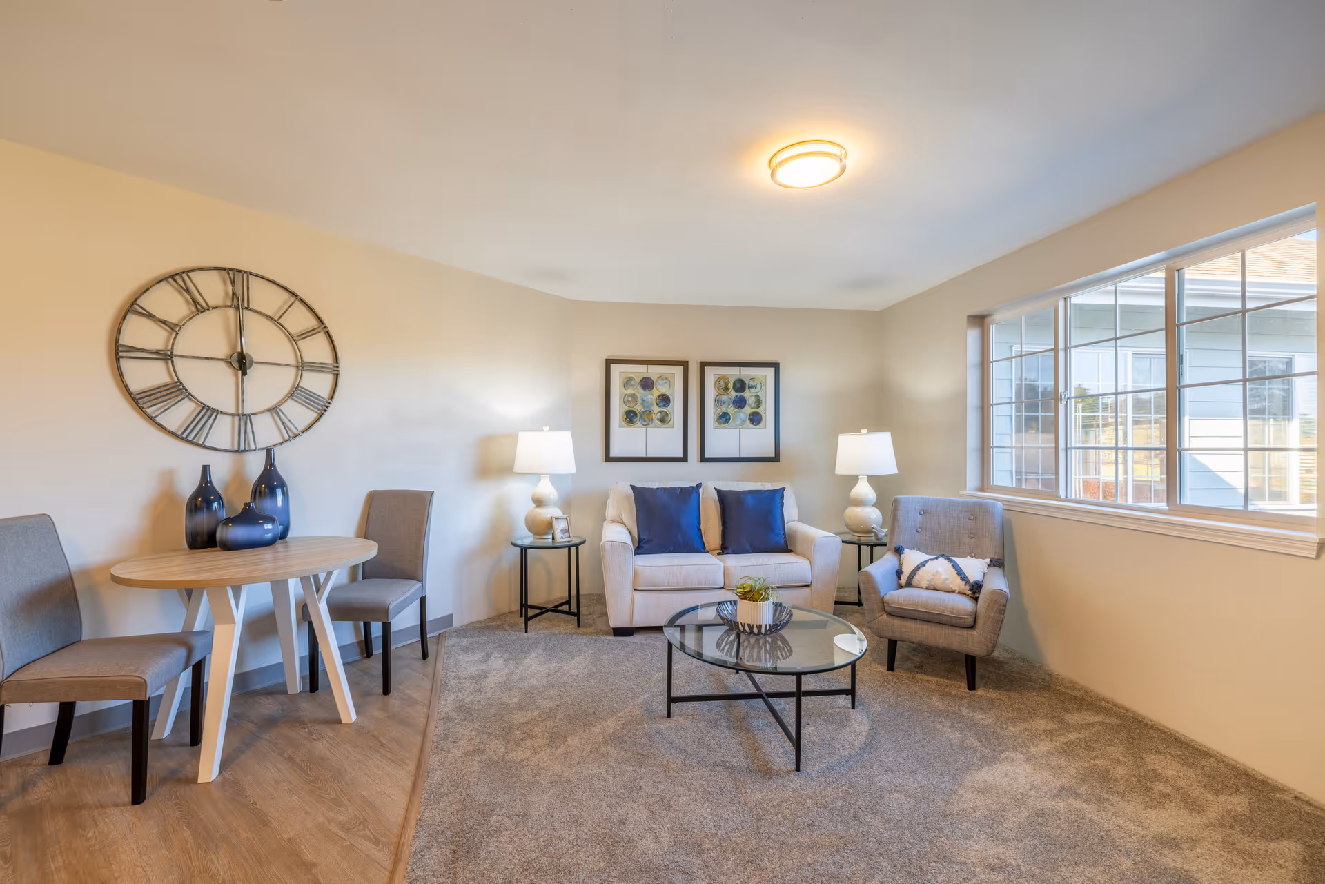 A cozy living room area with a beige loveseat adorned with two blue pillows, a gray armchair with a decorative pillow, and a round glass coffee table with a small plant on it. Two side tables with white lamps flank the loveseat. On the left side, there is a small wooden table with two gray chairs and three blue decorative vases. A large wall clock with Roman numerals hangs above the table. The room has beige walls, a large window letting in natural light, and a mix of carpet and wood flooring.