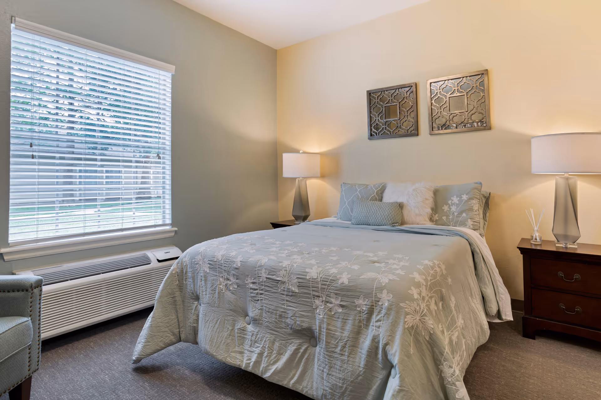 A cozy bedroom with a neatly made bed featuring light blue floral bedding and decorative pillows. There are two matching bedside tables with modern lamps on each side of the bed. Above the bed, two decorative mirrors hang on the beige wall. A window with white blinds lets in natural light, and a light blue armchair is partially visible next to the window.
