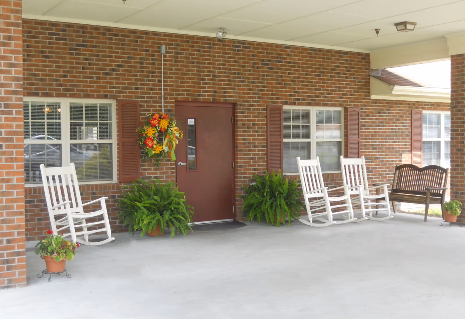 Covered outdoor patio area with a brick wall, a brown door, two windows with brown shutters, three white rocking chairs, a wooden bench, potted plants including ferns and flowers, and a hanging floral wreath on the wall.