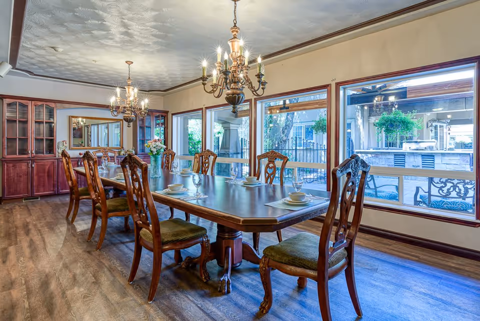 A spacious dining room with a long wooden table set with plates, bowls, and glasses. The room features ornate wooden chairs with green cushions, two elegant chandeliers hanging from a textured ceiling, large windows providing a view of an outdoor patio area with hanging plants, and a wooden cabinet with glass doors along one wall.