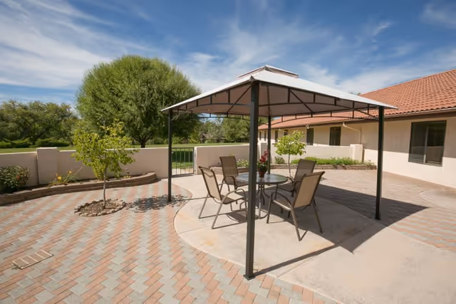 Outdoor patio area with a metal-framed gazebo covering a round glass table and four chairs. The patio is paved with interlocking bricks and surrounded by a low wall with small trees and plants. A building with a red-tiled roof and beige walls is visible in the background under a partly cloudy blue sky.
