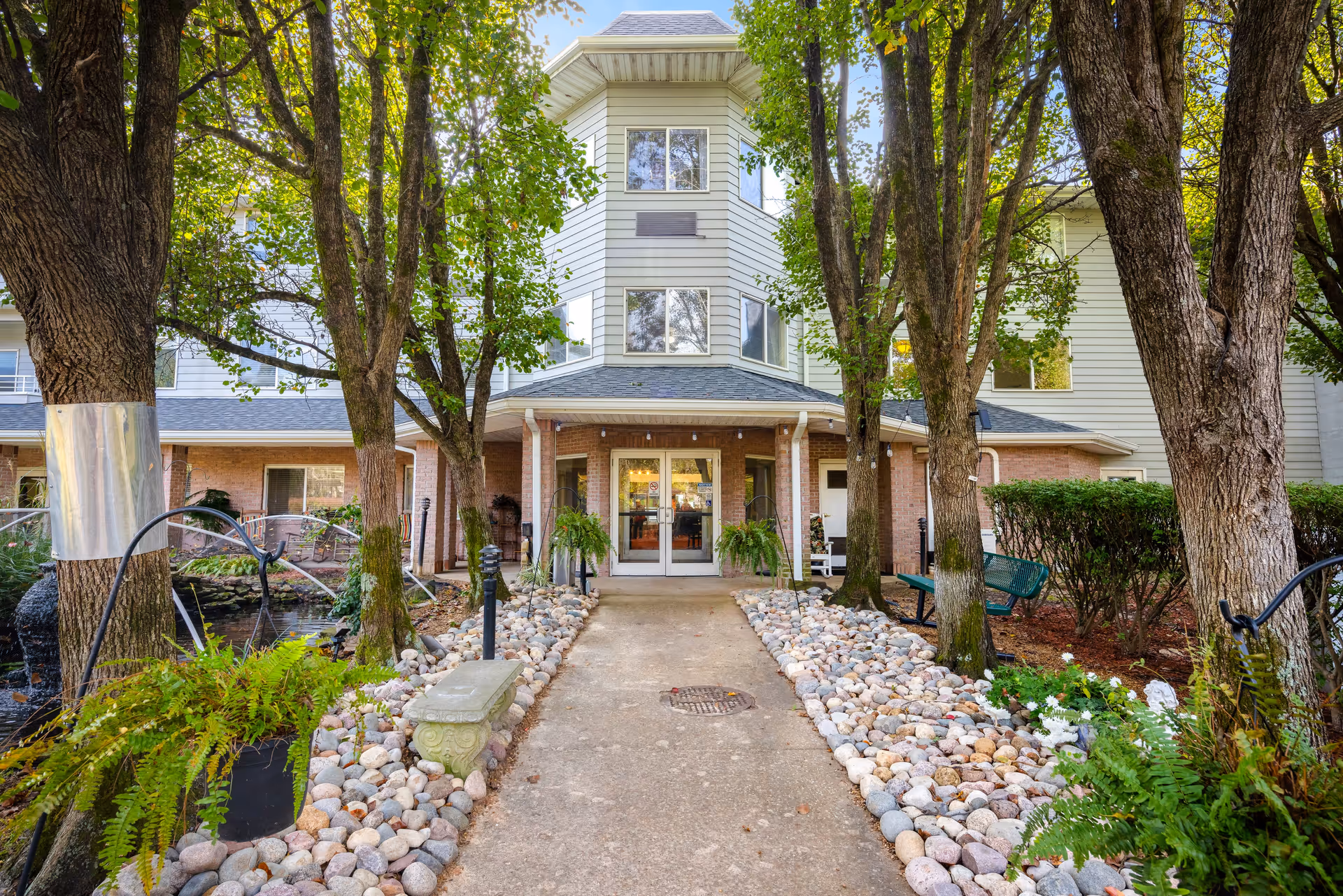 Entrance pathway to a senior living community building surrounded by trees and landscaped with rocks and plants. The building has a covered porch with double glass doors and multiple windows on the upper floors.