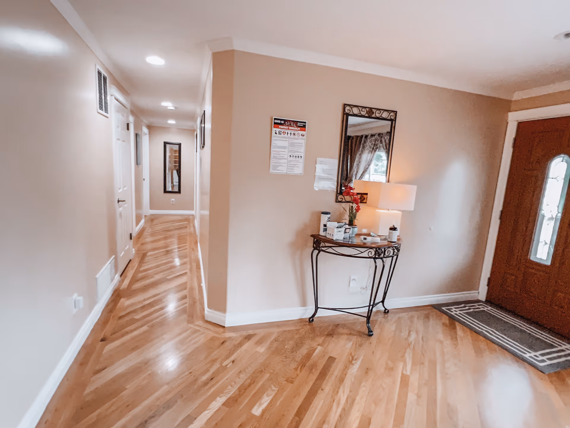 Interior hallway with polished wooden floors, beige walls, and white trim. A small decorative table with a lamp, flowers, and some items is placed against the wall near a wooden front door with a glass panel. A mirror hangs above the table, and a hallway with several doors and a wall mirror is visible in the background.