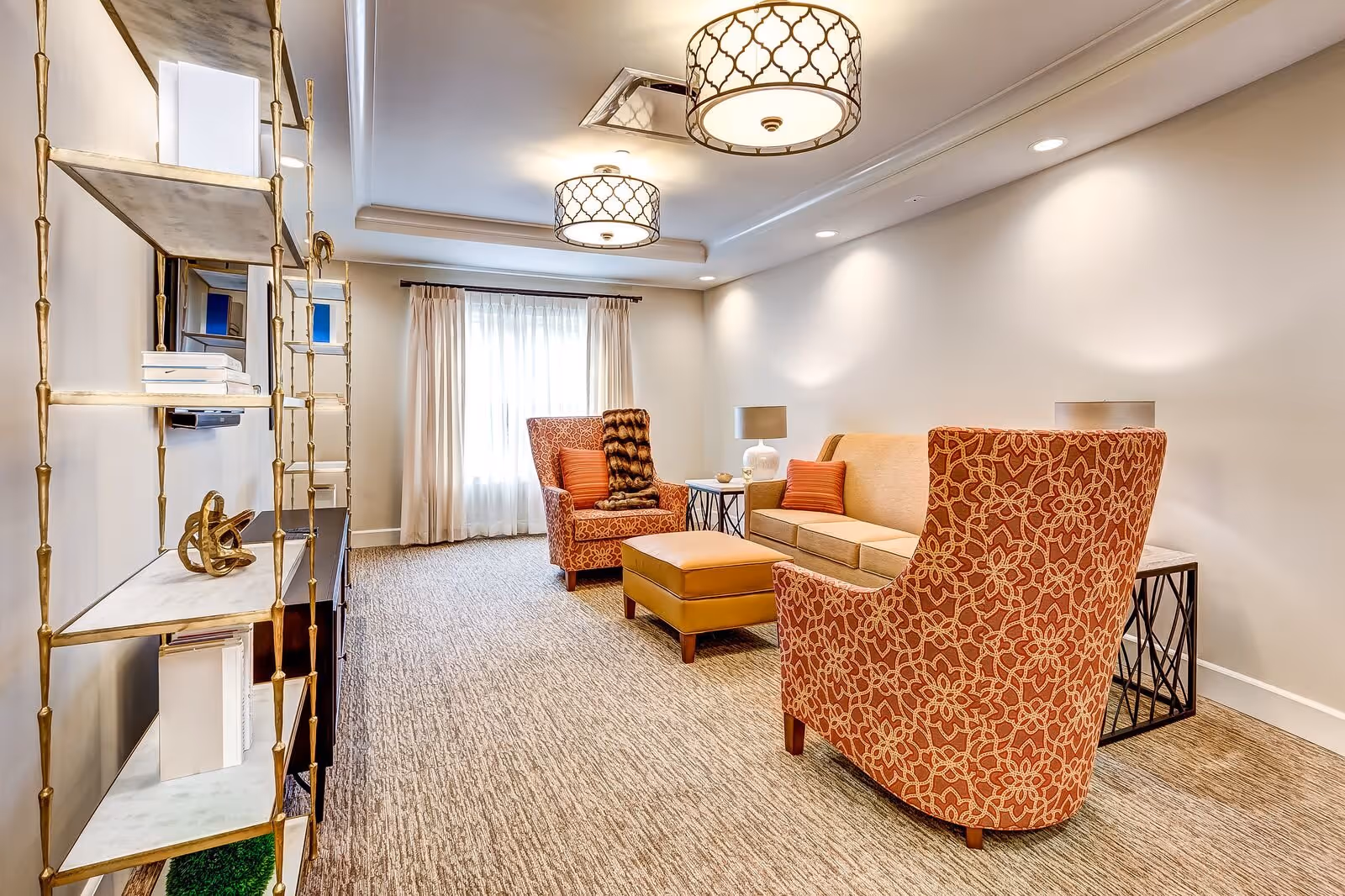 A well-lit living room with beige walls and carpeted floor featuring a beige sofa with orange pillows, two orange patterned armchairs, a matching ottoman, two side tables with lamps, a decorative shelving unit with books and ornaments, and two ceiling light fixtures. A window with sheer white curtains is at the far end of the room.