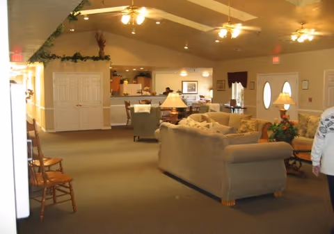 A spacious living room area in a senior living facility with beige sofas, armchairs, wooden chairs, and tables with lamps. The room has a high ceiling with ceiling fans and recessed lighting. There is a kitchen or dining area visible in the background, and the walls are decorated with plants and framed pictures. A person is partially visible on the right side of the image.