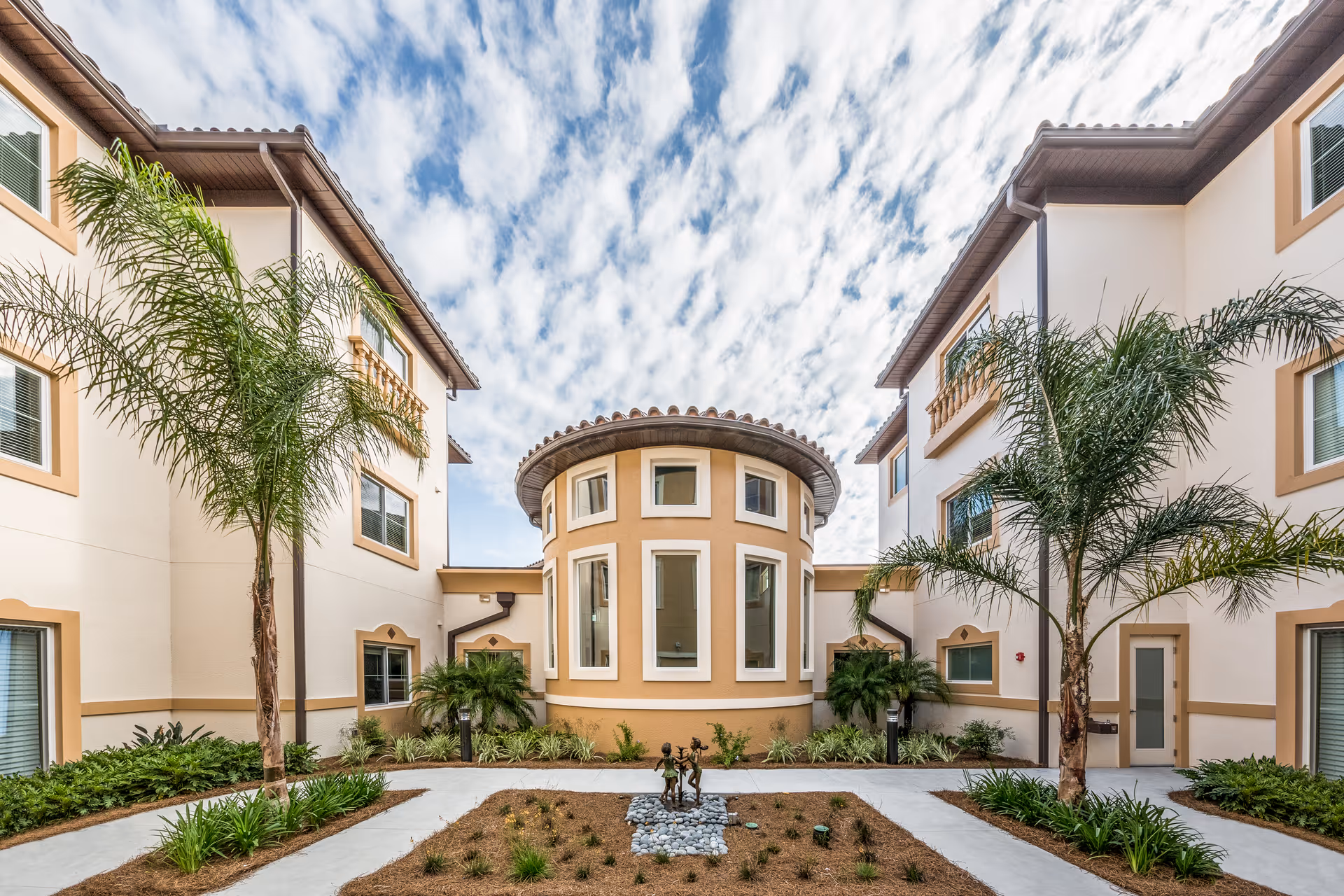Courtyard of a Mediterranean-style senior living complex with palm trees, walkways, landscaped beds and a central round building under a partly cloudy sky.