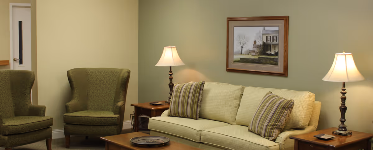 A cozy living room with a light green sofa adorned with striped pillows, two matching green armchairs, two wooden side tables each with a lamp, a wooden coffee table with a decorative tray, and a framed painting on the wall.