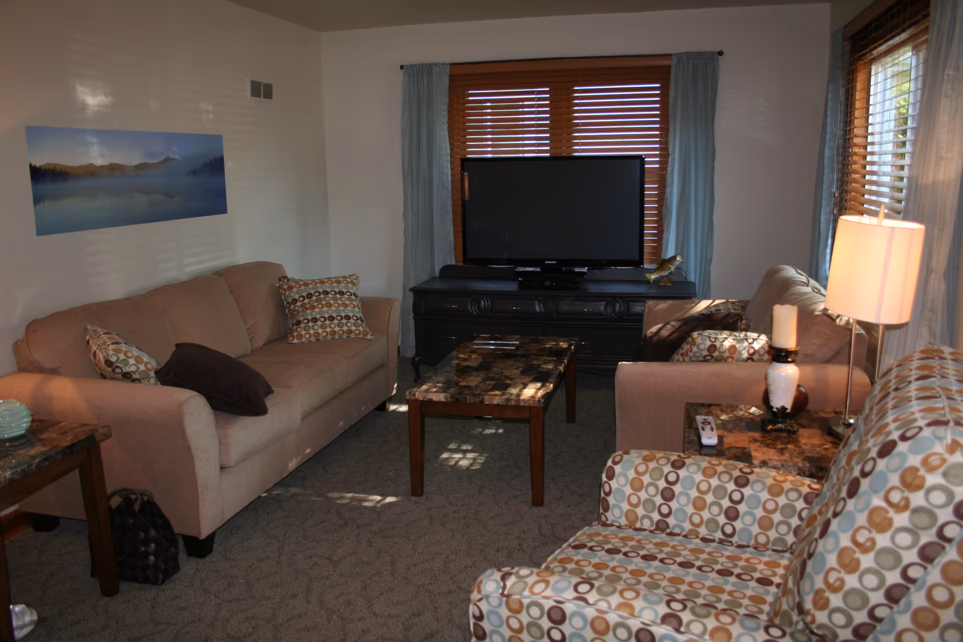 A cozy living room with two beige sofas, a patterned armchair, a coffee table, and a TV on a black stand in front of a window with wooden blinds and light blue curtains. There is a lamp and a candle on a side table next to the armchair, and a landscape painting on the wall.