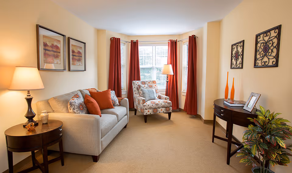 A cozy living room with a beige sofa adorned with patterned and solid orange cushions, a patterned armchair, and a wooden side table with a lamp. The room has beige walls, red curtains on the windows, two framed pictures on one wall, decorative wall art on another, and a small wooden console table with orange vases and a framed photo. There is also a potted plant on the floor near the console table.
