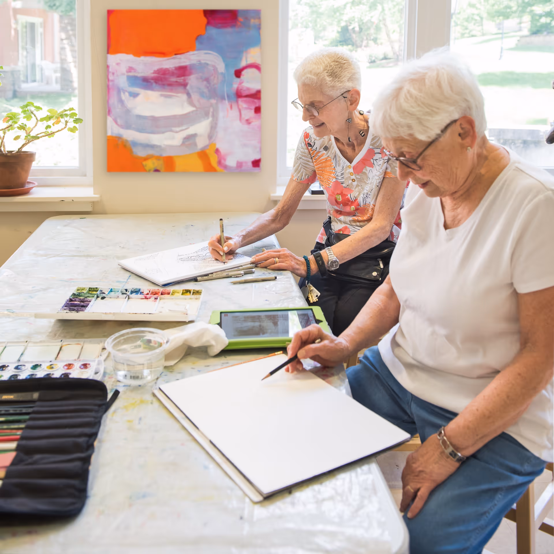 Two elderly women sitting at a table by a window, engaged in drawing and painting activities with art supplies including colored pencils, paint palettes, and sketchbooks. One woman is wearing a floral shirt and glasses, while the other is in a white shirt and glasses. A colorful abstract painting hangs on the wall behind them.