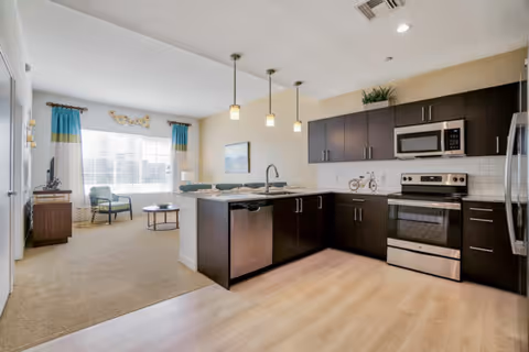 Open-plan living area with a modern kitchen featuring dark cabinetry, stainless-steel appliances, an island with pendant lights, and a carpeted sitting area by a large window.