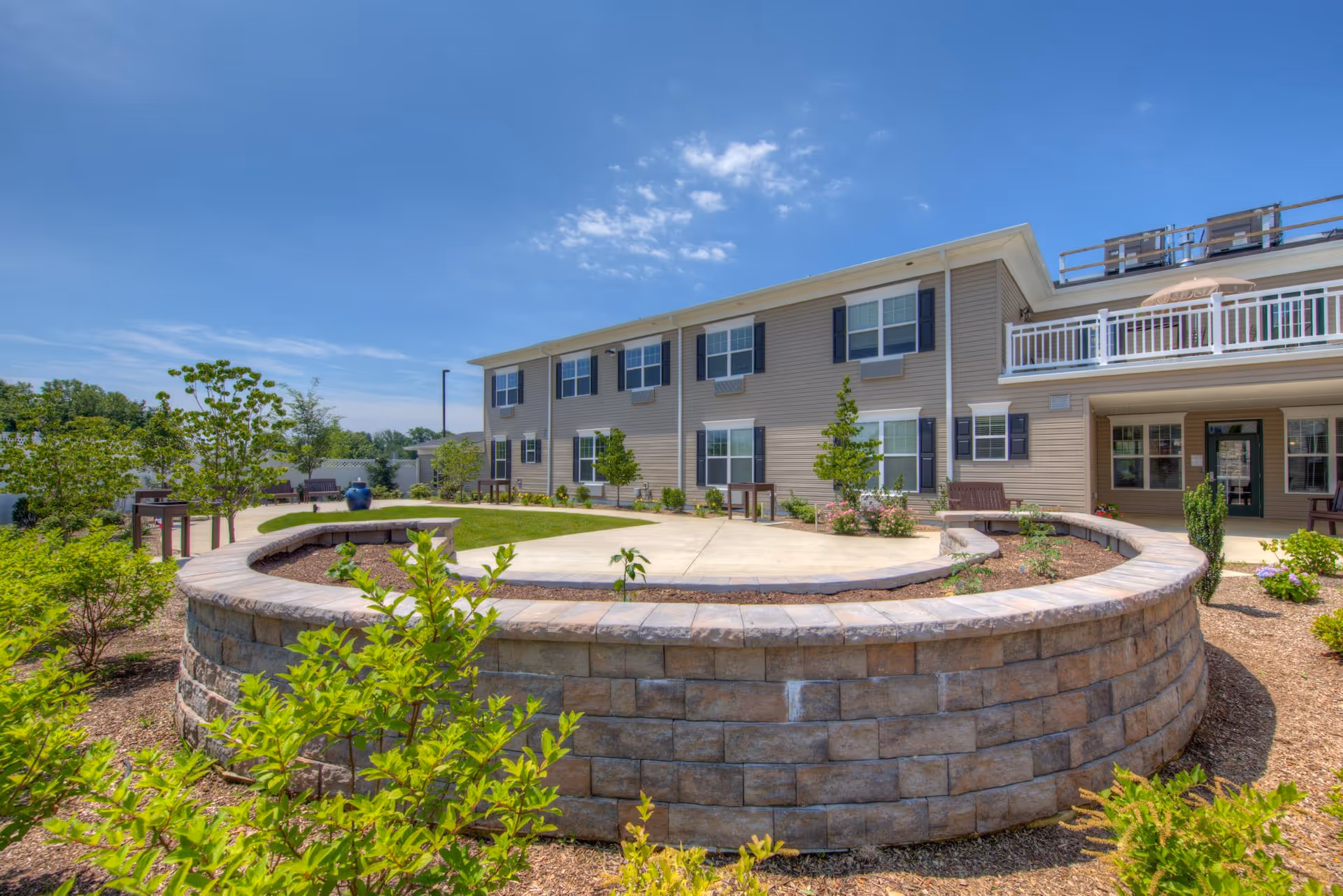 Outdoor courtyard area at Heather Glen At Ardenwoods featuring a circular stone retaining wall with a raised garden bed in the center, surrounded by small trees and shrubs. The two-story beige building with multiple windows and a balcony is visible in the background under a clear blue sky.