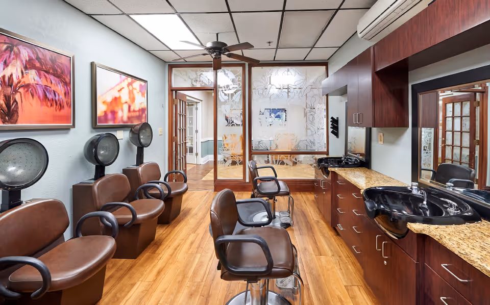Interior hair salon with multiple styling chairs and hooded dryers on the left and washbasins with mirrors and cabinetry on the right.