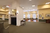 Spacious interior common area in an assisted living facility featuring a central white column with a fireplace, beige walls, carpeted floor, and several tables and chairs near large windows allowing natural light.