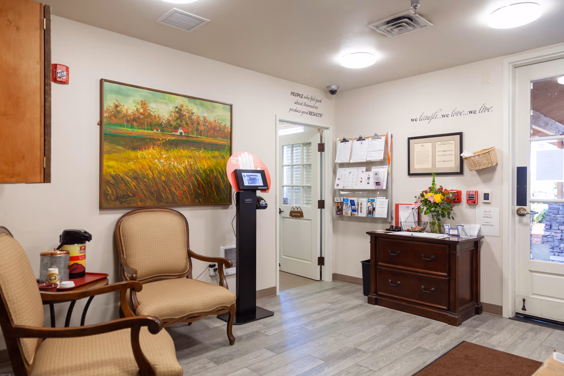 A cozy waiting area with two beige upholstered chairs and a small table holding a coffee dispenser and supplies. A large painting of a rural landscape with trees and a house hangs on the wall. There is a kiosk stand with a screen near an open doorway. On the right side, a wooden cabinet holds a vase of flowers and various papers. Above the cabinet, wall decals read 'we laugh... we love... we live.' and 'PEOPLE who feel good about themselves produce good RESULTS!'. A glass door leads outside, showing part of a stone wall and wooden beams.