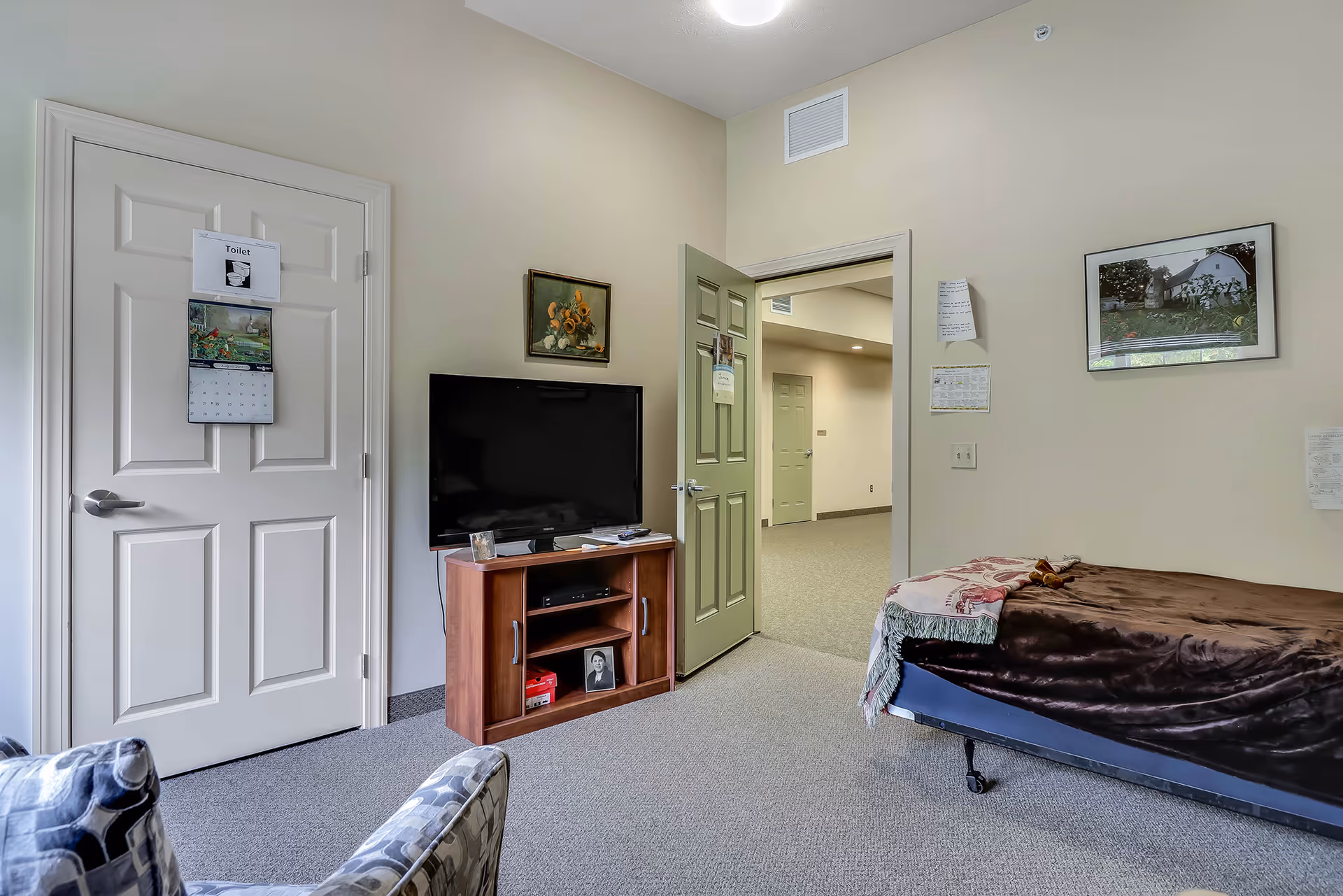 A cozy assisted living room with a single bed covered in a brown blanket and a floral throw, a flat-screen TV on a wooden stand, a patterned armchair, and two doors, one labeled 'Toilet'. The walls are decorated with framed pictures and papers.