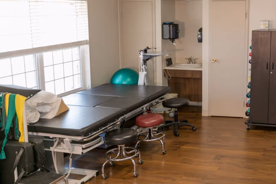 Physical therapy/rehab room with an adjustable treatment table, several rolling stools, an exercise ball, sink, and storage cabinet.