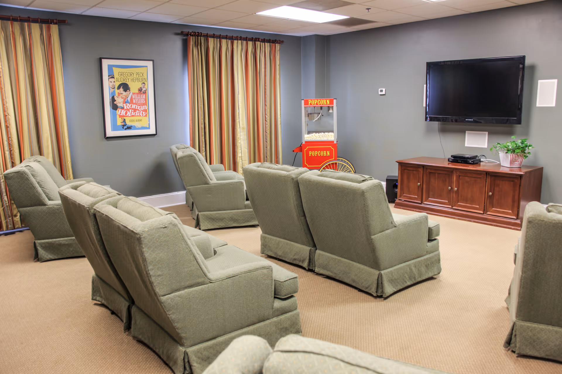 A cozy media room with multiple green upholstered recliner chairs arranged in rows facing a wall-mounted flat screen TV. There is a wooden cabinet below the TV with a plant on top, and a vintage-style popcorn machine in the corner. The walls are painted gray with striped curtains covering the windows and a framed movie poster on the wall.