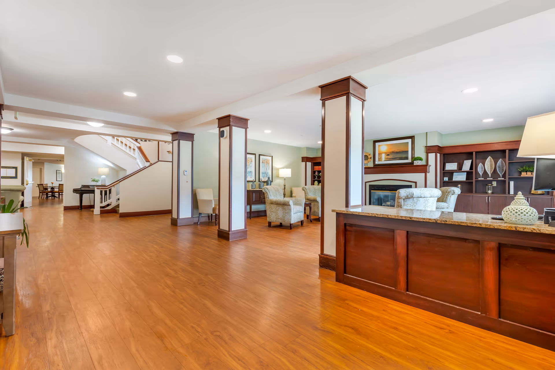 Spacious senior living facility common area with wooden flooring, a reception desk on the right, comfortable armchairs arranged near a fireplace, decorative plants, framed artwork on the walls, and a staircase leading to the upper floor.