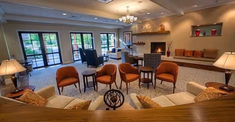 Bright, comfortable lounge with multiple armchairs and sofas arranged around small tables, a fireplace, and large glass doors leading to an outdoor patio.