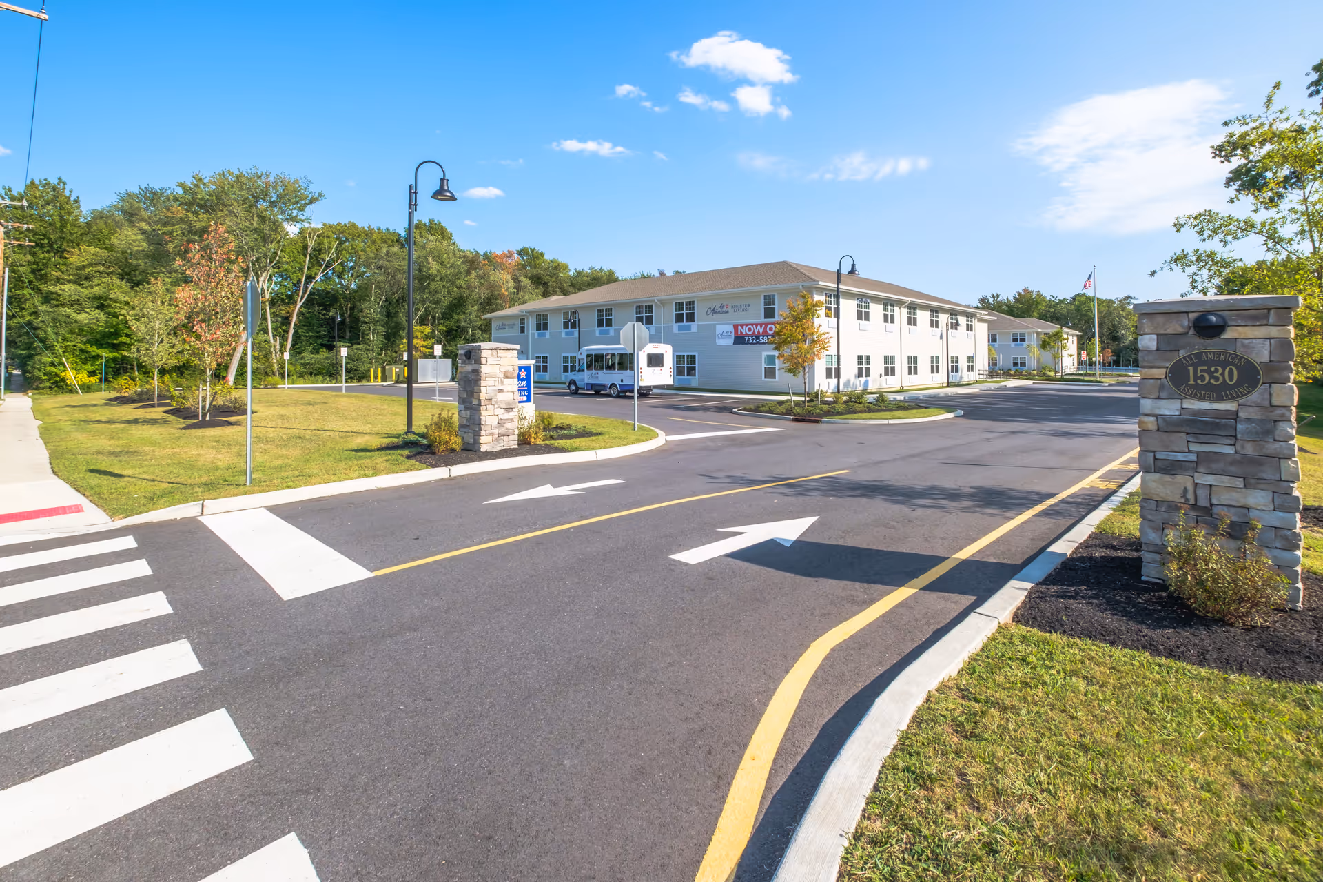 Exterior view of All American Assisted Living at Tinton Falls facility showing a two-story building with multiple windows, a driveway with directional arrows, landscaped grass and trees, stone pillars with signage, and a clear blue sky.