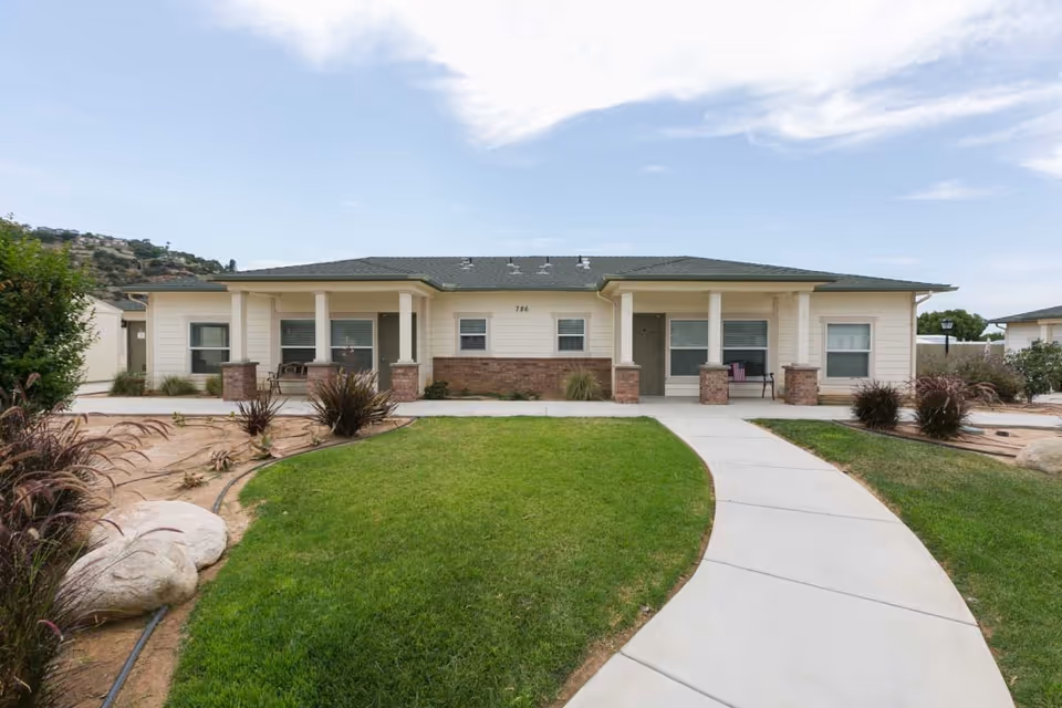 Front exterior view of a single-story senior living facility building with a green lawn, a curved concrete walkway leading to the entrance, and some landscaping with rocks and plants. The building has a light-colored facade with brick accents and multiple windows.