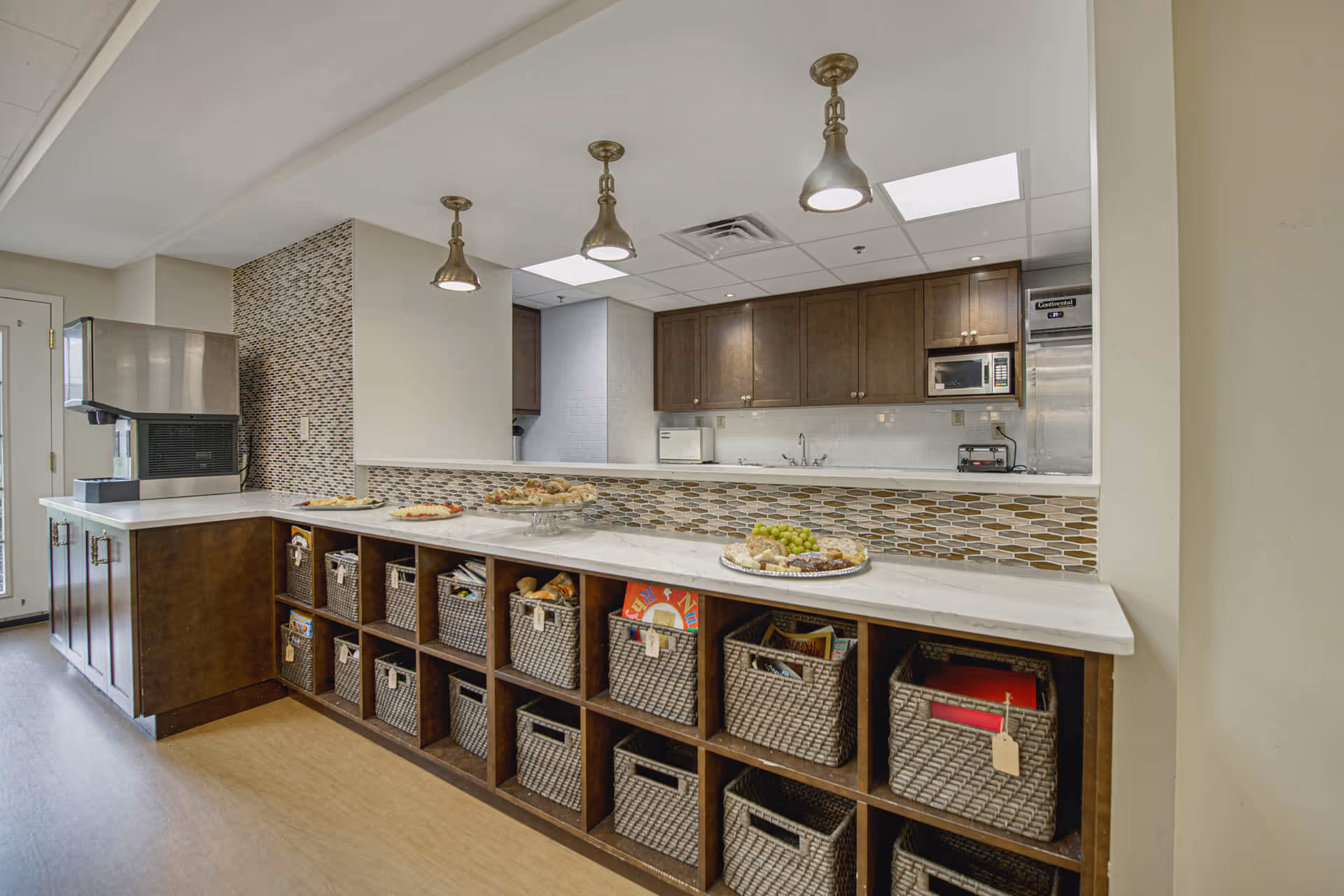 A kitchen area in a senior living facility with a long counter featuring a white countertop and multiple woven baskets stored underneath. The back wall has dark wooden cabinets, a microwave, a toaster, and a refrigerator. There are three pendant lights hanging from the ceiling, and the backsplash is decorated with a mosaic tile pattern. Plates with assorted food items are placed on the counter.