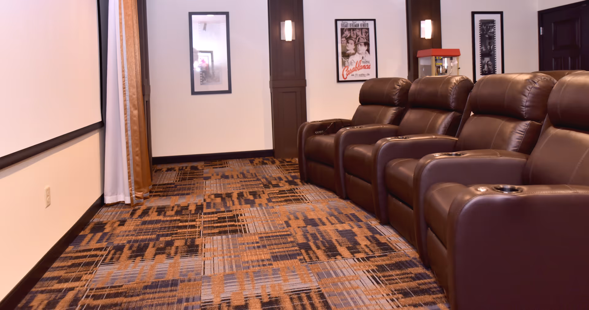 A small private theater room with four brown leather recliner chairs arranged in a row facing a large white projection screen. The room has patterned carpet flooring, framed movie posters on the walls, and a popcorn machine in the corner.