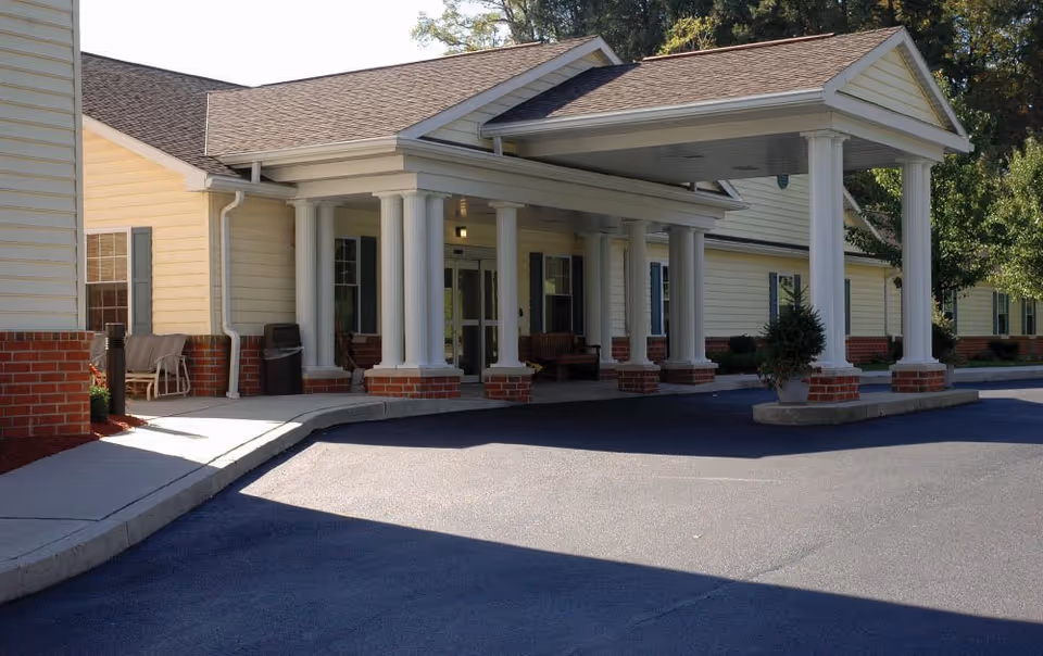Entrance of a senior living facility with a covered drop-off area supported by white columns. The building exterior features light yellow siding with red brick accents and several windows. There are benches and a trash can near the entrance, and some greenery is visible around the building.