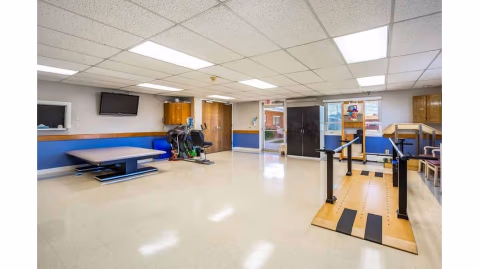 A spacious rehabilitation room with exercise equipment including a stationary bike, parallel bars for walking practice, a therapy table, a wall-mounted TV, and a small basketball hoop. The room has a tiled floor, white ceiling with fluorescent lights, and blue and white walls. There are windows and a door leading outside.