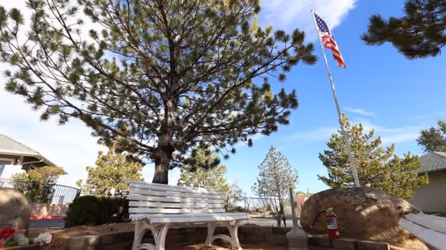 Outdoor seating area with a white bench under a large pine tree, an American flag on a tall flagpole, large rocks, and a clear blue sky with some clouds.